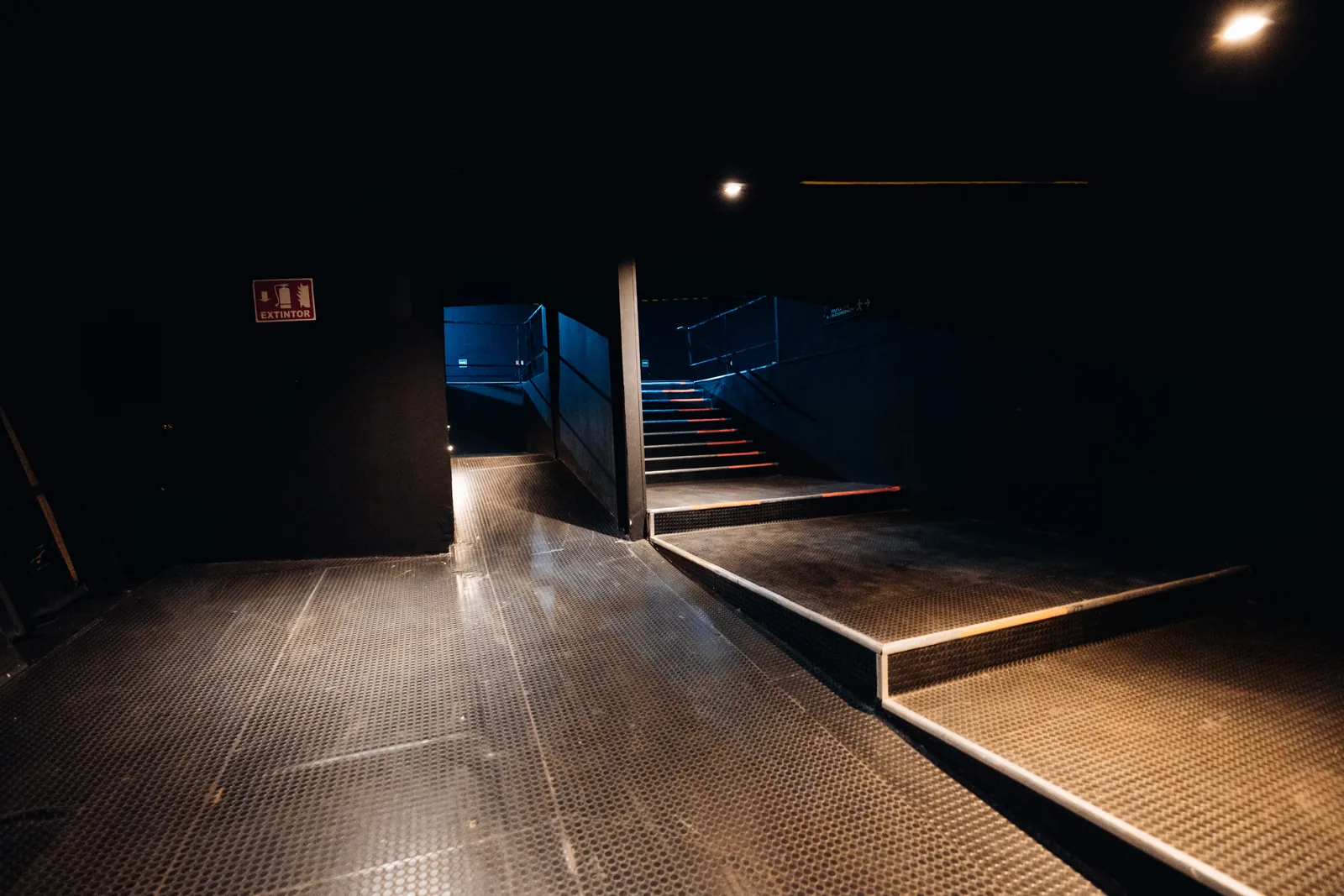 Pasillo oscuro con piso metálico, señal de extintor y escaleras iluminadas al fondo.