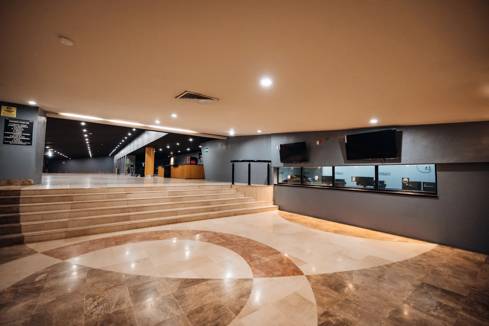 Lobby con escaleras, piso de mármol con diseño circular y área de mostradores con pantallas negras.
