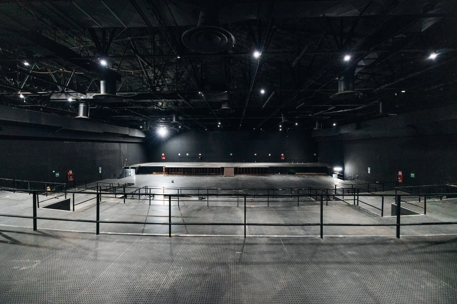 Gran auditorio vacío con escenario amplio y barandales negros bajo techo con luces y ductos.
