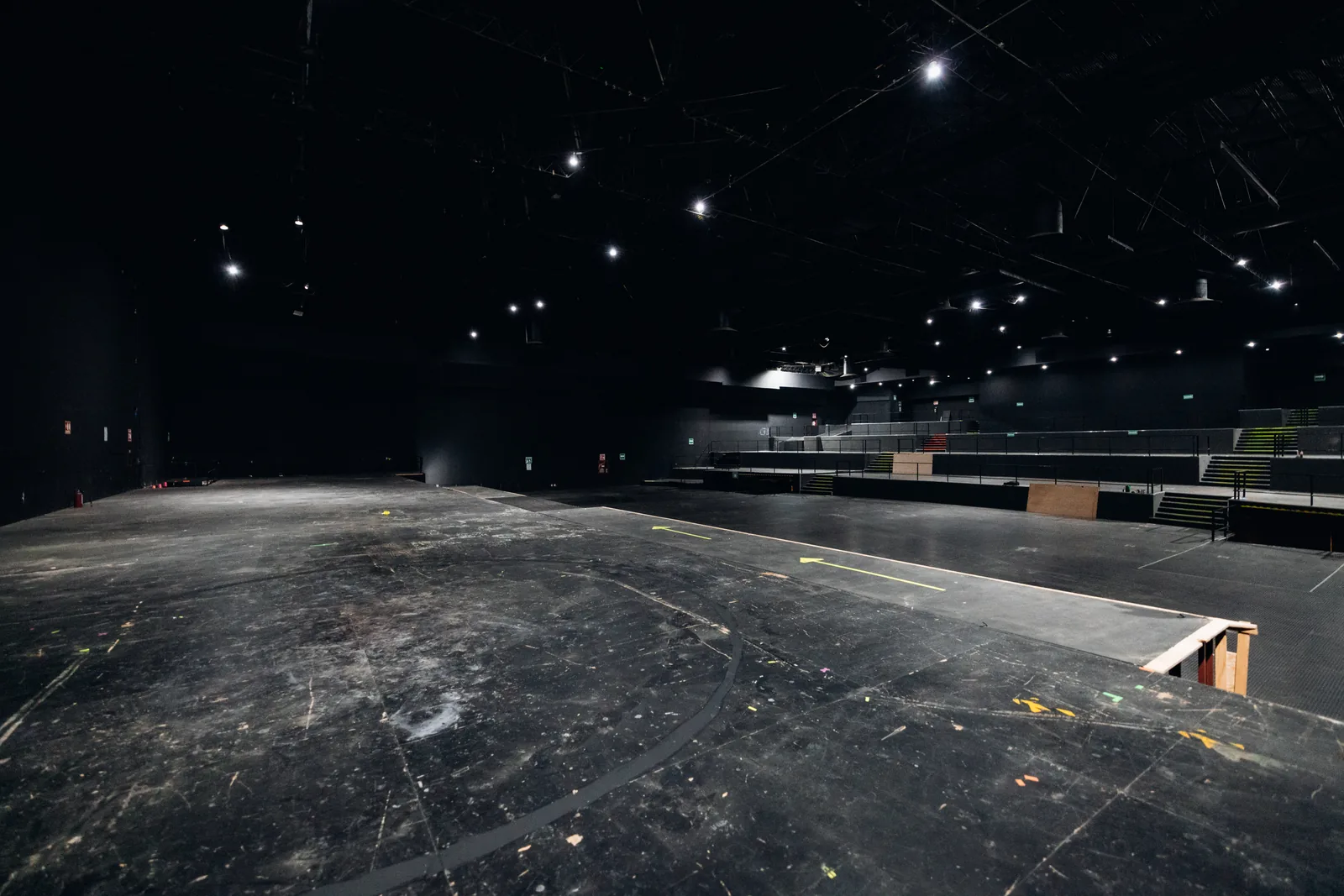Escenario amplio y vacío con iluminación en techo y gradas al fondo en un foro negro.