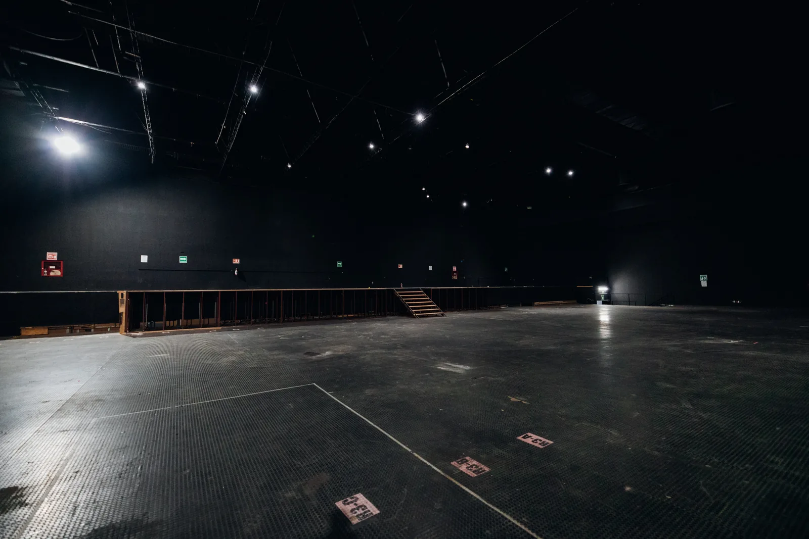 Espacio grande y vacío con piso negro y paredes oscuras iluminado con luces blancas en el techo.